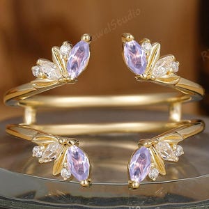 May include: A gold-coloured ring guard with four decorative sections, each featuring a lavender-coloured gemstone and smaller clear stones. The ring guard is designed to enhance the look of a ring. The ring is displayed on a decorative surface.