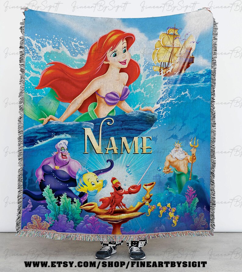 May include: A woven throw blanket featuring a colorful illustration of Ariel from The Little Mermaid. The design includes Ariel, Ursula, King Triton, and other characters, with the word "NAME" in the center. The blanket has a fringed edge.