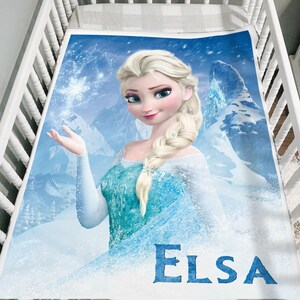 Puede incluir: Una manta para beb&eacute; con un gr&aacute;fico de Elsa de la pel&iacute;cula Frozen. La manta tiene una combinaci&oacute;n de colores azul y blanco con un fondo de monta&ntilde;as nevadas. El nombre "Elsa" est&aacute; impreso en azul en la parte inferior.