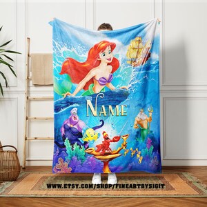 May include: A blue blanket featuring Ariel from The Little Mermaid. The blanket has a vibrant illustration of Ariel, Ursula, Flounder, Sebastian, King Triton, and a ship. The word "Name" is written in a cursive font. The blanket is a cozy home decor item.
