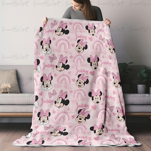 K&ouml;nnte beinhalten: Eine rosa Decke mit einem sich wiederholenden Muster von Minnie Mouse, Schleifen, Regenbogen, Herzen und Sternen. Der Name "Jenny" ist in Schreibschrift geschrieben. Die Decke wird von einer Person gehalten.