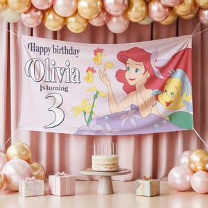 May include: A pink banner with the text "Happy birthday Olivia Is turning 3" and a cartoon illustration of Ariel and Flounder from The Little Mermaid. The banner is decorated with balloons and a birthday cake.