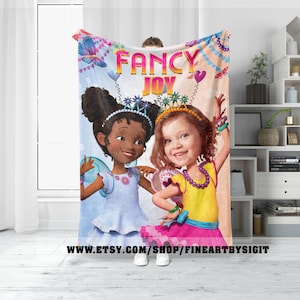 May include: A pink and blue fleece blanket with two cartoon girls wearing fairy wings and crowns. The text "FANCY JOY" is printed on the blanket.