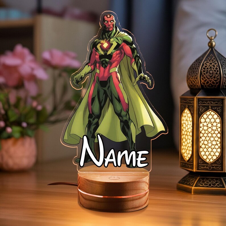 May include: A superhero-themed night light featuring a character in red, green, and yellow. The character is standing on a wooden base with the word "Name" below. The lamp is plugged in and lit, with a warm glow. A decorative lantern is in the background.