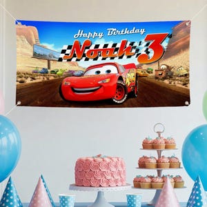 May include: A banner with the text "Happy Birthday Noah 3" featuring the character Lightning McQueen from the movie Cars. The banner is blue with a desert landscape background. Party decorations include a pink cake, cupcakes, and balloons.