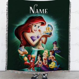 May include: A woven throw blanket featuring Ariel from The Little Mermaid. The design includes Ariel, King Triton, Sebastian, Flounder, and other characters. The blanket has a dark green background with the word "NAME" at the top. The blanket has a fringed edge.