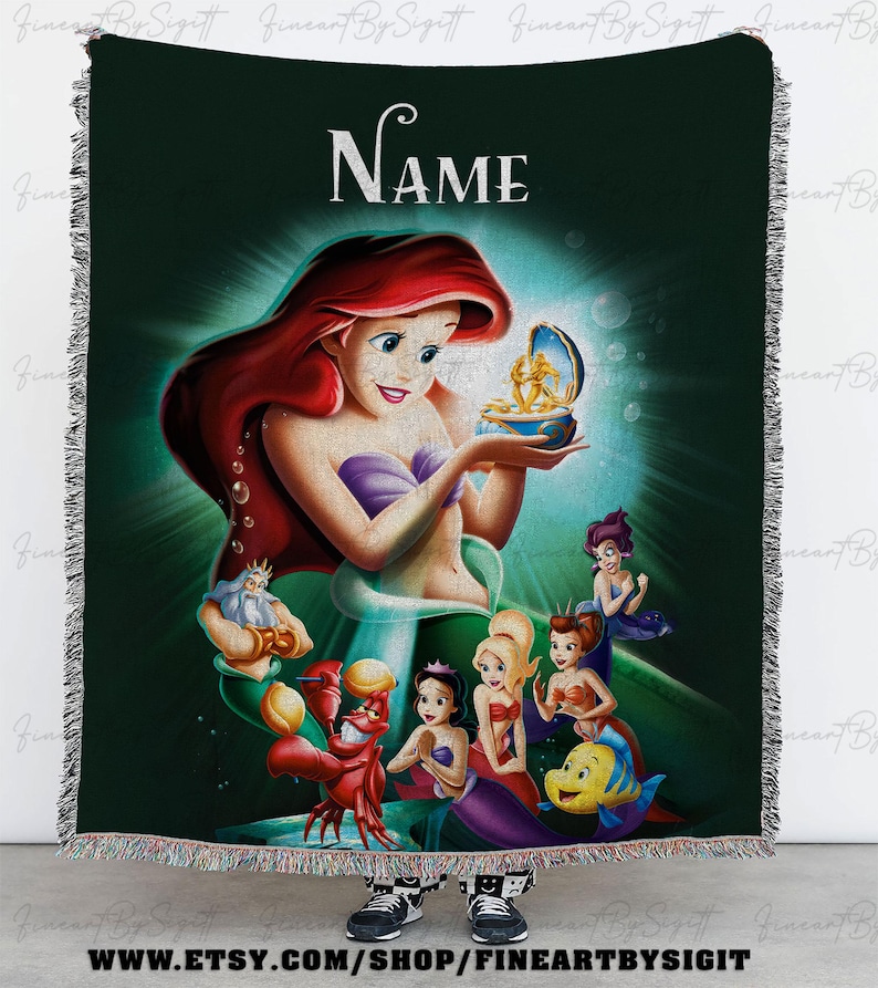 May include: A woven throw blanket featuring Ariel from The Little Mermaid. The design includes Ariel, King Triton, Sebastian, Flounder, and other characters. The blanket has a dark green background with the word "NAME" at the top. The blanket has a fringed edge.