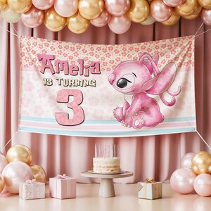 May include: A pink banner with the text "Amelia is turning 3" and a cartoon character. The banner is decorated with floral patterns and pastel stripes. A small cake with candles sits on a pedestal table, surrounded by balloons and gifts.