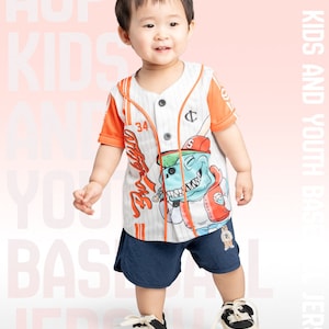 May include: A young child wearing a white and orange baseball jersey with a cartoon dinosaur graphic. The jersey has a button-up front and a striped pattern. The child is also wearing navy blue shorts and black sneakers.