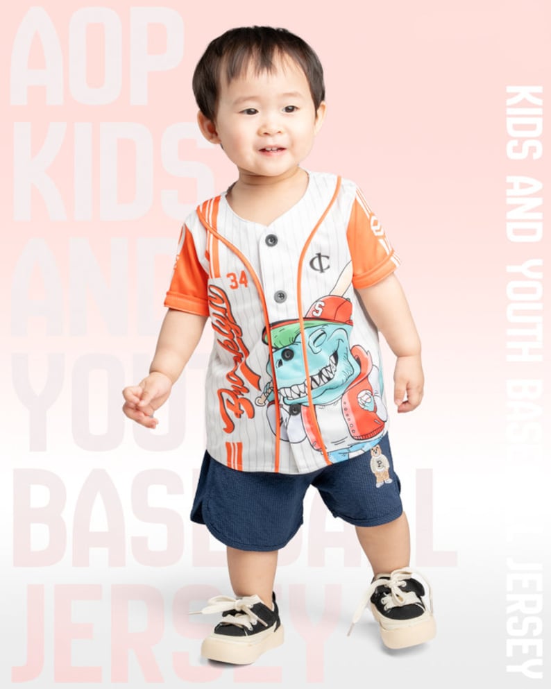 May include: A young child wearing a white and orange baseball jersey with a cartoon dinosaur graphic. The jersey has a button-up front and a striped pattern. The child is also wearing navy blue shorts and black sneakers.