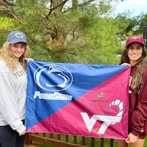 May include: Two people holding a Penn State and Virginia Tech flag. The flag is blue and pink with the Penn State Nittany Lion logo on the blue side and the Virginia Tech Hokie Bird logo on the pink side. The text "Penn State" and "VT" are on the flag.