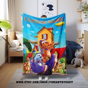 May include: A blue fleece blanket featuring cartoon characters and a house illustration. The blanket has the text "CUSTOM NAME" in orange. The bottom of the blanket has the website address www.etsy.com/shop/fineartbysigit.