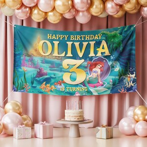 May include: A birthday banner with the text "Happy Birthday Olivia 3 is Turning" featuring an underwater scene with a mermaid. The banner is decorated with balloons, gifts, and a cake with candles, creating a festive atmosphere.