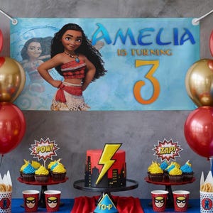 K&ouml;nnte beinhalten: Eine Geburtstagsparty-Szene mit einem Banner mit einer Zeichentrickfigur und dem Text "AMELIA WIRD 3". Der Tisch ist mit einem Kuchen, Cupcakes, Luftballons und Tassen im Superhelden-Stil dekoriert. Der Kuchen ist rot mit einem gelben Blitz.