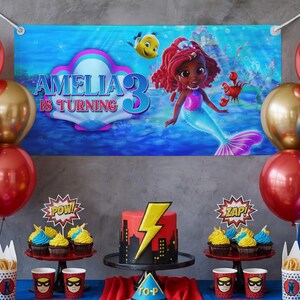 May include: A birthday party scene with a banner that reads "AMELIA IS TURNING 3". The banner features a mermaid illustration. The table has a cake with a lightning bolt design, cupcakes, and superhero-themed cups and decorations. Balloons in red and gold complete the festive look.