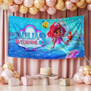 May include: A blue banner with the text "AMELIA 3 IS TURNING" features a mermaid illustration. The banner is decorated with a cartoon fish and crab. The scene is set against a backdrop of balloons, a cake, and gifts, creating a festive birthday atmosphere.