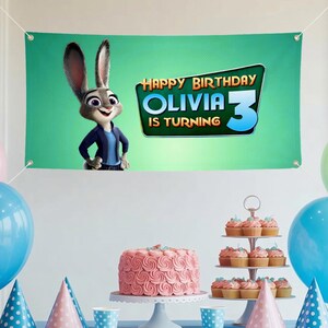 Puede incluir: Una pancarta verde con el texto "HAPPY BIRTHDAY OLIVIA 3 IS TURNING" y un personaje de dibujos animados. Se exhiben un pastel rosa y cupcakes, junto con globos y gorros de fiesta azules y rosas. La pancarta es una decoraci&oacute;n de fiesta de cumplea&ntilde;os.