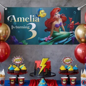 May include: A birthday party scene with a banner featuring Ariel from The Little Mermaid. The banner reads "Amelia Is turning 3." A red and yellow cake with a lightning bolt design sits on a black cake stand. Cupcakes, balloons, and themed cups complete the festive setup.