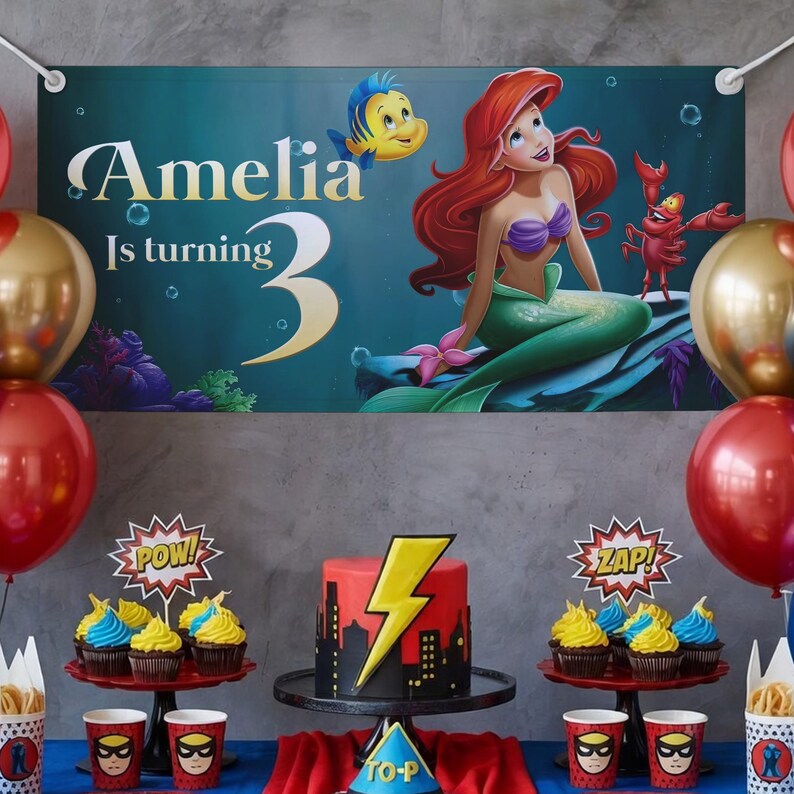 May include: A birthday party scene with a banner featuring Ariel from The Little Mermaid. The banner reads "Amelia Is turning 3." A red and yellow cake with a lightning bolt design sits on a black cake stand. Cupcakes, balloons, and themed cups complete the festive setup.