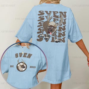 May include: Light blue t-shirt featuring a cartoon reindeer named Sven. The back of the shirt has the name "SVEN" in a repeating pattern with a reindeer graphic. The front has a smaller Sven graphic with "EST. 2013".