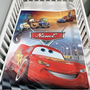 May include: A colorful crib sheet featuring characters from the animated movie "Cars." The design includes Lightning McQueen, Mater, and other vehicles against a desert landscape and race track background. The sheet has the word "Name" on it.