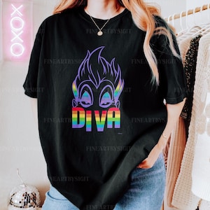 Op de afbeelding: Zwart T-shirt met een paars cartoonfiguur met regenboogtekst die "Diva" zegt.