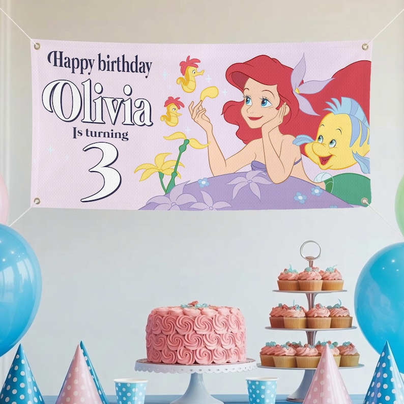 May include: A banner with the text "Happy birthday Olivia Is turning 3" featuring a cartoon illustration of Ariel and Flounder from The Little Mermaid. The banner is pink and is part of a birthday party setup.