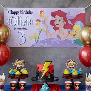 May include: A birthday party scene with a banner that reads "Happy birthday Olivia Is turning 3". The banner features a cartoon of Ariel from The Little Mermaid. The table has a cake, cupcakes, and superhero-themed decorations.