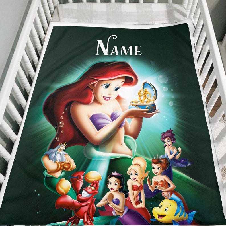 May include: A green blanket featuring Ariel from The Little Mermaid, holding a treasure box. Other characters from the movie are also depicted. The word "Name" is written in white script at the top. The blanket is placed in a white crib.