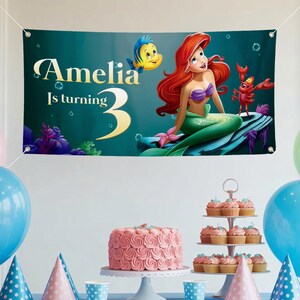 May include: A banner featuring Ariel from The Little Mermaid, with the text "Amelia Is turning 3." The banner has a teal background with illustrations of Flounder and Sebastian. A pink cake, cupcakes, and balloons are in the foreground.