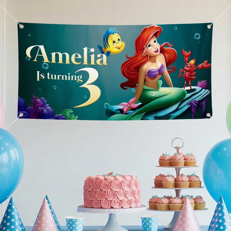 May include: A banner featuring Ariel from The Little Mermaid, with the text "Amelia Is turning 3." The banner has a teal background with illustrations of Flounder and Sebastian. A pink cake, cupcakes, and balloons are in the foreground.