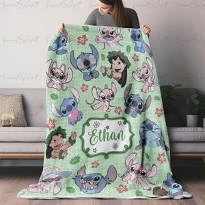 May include: A light green blanket featuring cartoon characters, including Stitch and Lilo, with a grid pattern and floral accents. The blanket has the name "Ethan" in a decorative frame. The blanket is held up by a person.
