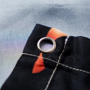 May include: Close-up of a black fabric corner with a silver grommet. The fabric has an orange and red pattern and is stitched with white thread. The background is a light blue and purple gradient.