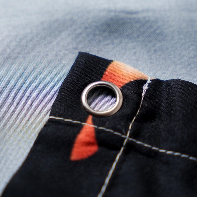 May include: Close-up of a black fabric corner with a silver grommet. The fabric has an orange and red pattern and is stitched with white thread. The background is a light blue and purple gradient.