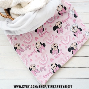 K&ouml;nnte beinhalten: Eine rosa Babydecke mit einem Muster aus Minnie Mouse, Schleifen, Regenbogen und dem Namen "Jenny". Die Decke hat ein wei&szlig;es Sherpa-Futter und ist teilweise in einem Weidenkorb gefaltet. Die Decke ist aus weichem Material.