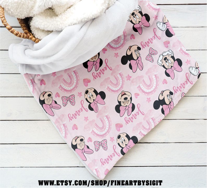 K&ouml;nnte beinhalten: Eine rosa Babydecke mit einem Muster aus Minnie Mouse, Schleifen, Regenbogen und dem Namen "Jenny". Die Decke hat ein wei&szlig;es Sherpa-Futter und ist teilweise in einem Weidenkorb gefaltet. Die Decke ist aus weichem Material.