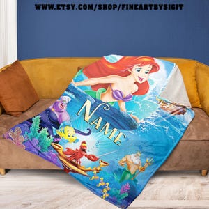 May include: A blue and white throw blanket featuring Ariel from The Little Mermaid. The blanket has a personalized name in gold lettering. Other characters include Flounder, Sebastian, Ursula, and King Triton. The blanket is displayed on a brown couch.