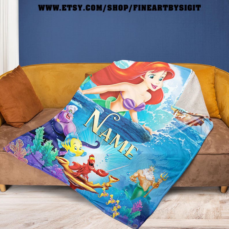 May include: A blue and white throw blanket featuring Ariel from The Little Mermaid. The blanket has a personalized name in gold lettering. Other characters include Flounder, Sebastian, Ursula, and King Triton. The blanket is displayed on a brown couch.