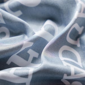 May include: Close-up of a light blue fabric with white lettering. The fabric is draped, creating soft folds and shadows. The white letters are large and stylized, contrasting against the blue background. The overall texture appears smooth and possibly silky.