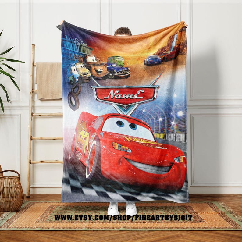 May include: A Cars-themed blanket featuring Lightning McQueen in a race scene. The blanket has a vibrant design with the text "Name" and other Cars characters. The background includes a checkered flag and a tunnel.