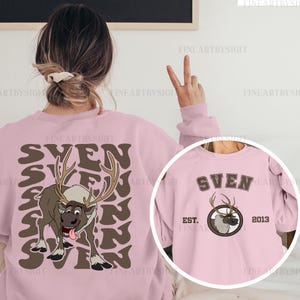 May include: Pink sweatshirt featuring a cartoon reindeer with the name "SVEN" in brown lettering. The design includes a circular graphic with the reindeer and the text "EST. 2013". The sweatshirt is a light pink color.