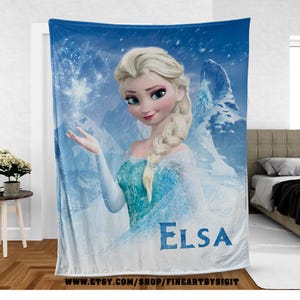 Puede incluir: Una manta azul con el personaje animado Elsa de la pel&iacute;cula Frozen. Elsa est&aacute; representada con cabello rubio, un vestido azul y un copo de nieve. La palabra "ELSA" est&aacute; impresa en azul en la parte inferior de la manta.