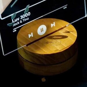 May include: A wooden, circular music player with a play/pause button and forward/backward controls. The text "Luv 3000 Jack & Tina" is visible on a clear acrylic panel above the player. The player is a warm, golden color.