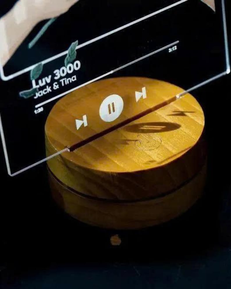 May include: A wooden, circular music player with a play/pause button and forward/backward controls. The text "Luv 3000 Jack & Tina" is visible on a clear acrylic panel above the player. The player is a warm, golden color.