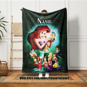 May include: A dark green fleece blanket featuring a colorful illustration of Ariel from The Little Mermaid. The image includes Ariel holding a treasure, with other characters from the movie. The word "Name" is at the top.