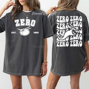 Puede incluir: Una camiseta gris oscuro con un gr&aacute;fico blanco de un fantasma con el texto "ZERO" en una fuente retro. La parte posterior de la camiseta tiene el texto "ZERO ZERO" repetido en una fuente retro con el gr&aacute;fico del fantasma en el centro.