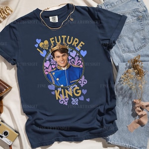 May include: Navy blue t-shirt featuring a graphic of a young man wearing a crown, with the words "I the FUTURE KING" in gold glitter. The design includes purple and blue hearts and fleur-de-lis.