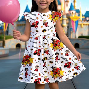 May include: A white dress featuring a pattern of characters and the text "The Incredibles" in black. The dress has a flared skirt and short sleeves. The design includes red, black, yellow, and orange elements.