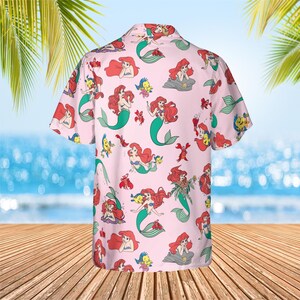 May include: A short-sleeve button-down shirt in light pink featuring an all-over repeating print of Ariel the mermaid and Flounder the fish from The Little Mermaid. The shirt has a classic collar and a buttoned front placket with a straight hem.