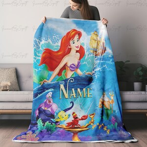 May include: A blue blanket featuring Ariel from The Little Mermaid. The design includes Ariel, Ursula, Sebastian, Flounder, King Triton, and a ship. The word "NAME" is also visible on the blanket.